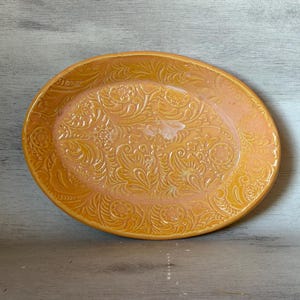 Op de afbeelding: Ovale keramische serveerschaal in een warme geel-oranje tint. De schaal heeft een reliëf bloemen- en krullenontwerp over het oppervlak. De randen zijn licht verhoogd en de algehele esthetiek is rustiek en decoratief.