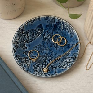 Puede incluir: Un plato de cerámica azul con una escena submarina con tortugas y peces. El plato contiene anillos de oro y un collar con un colgante de sol. Se ve un libro azul con el texto "THE COAST".
