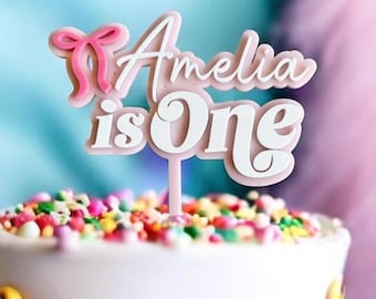May include: Pink and white cake topper with the text "Amelia is One" and a pink bow.