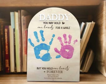 May include: Wooden sign with a handprint design and the text "Daddy You May Hold Our Hands For A While But You Hold Our Hearts Forever Happy Father's Day We Love You Dad Lots of Love [Name] & [Name] xx"