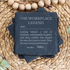 May include: A square black slate coaster featuring the engraved text THE WORKPLACE LEGEND, noun, followed by a definition about leaving a trail of kindness, laughter, and wisdom, ending with see also: Helen, with rough, textured edges.