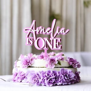 May include: Pink cake topper with the text "Amelia is One" in white lettering. The topper is on a two-tiered cake decorated with purple flowers.