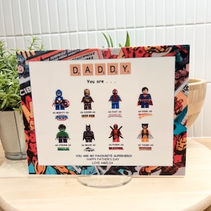 May include: A white card with a comic book background featuring a Lego figure of each superhero. The card reads "DADDY You are..." and lists the superheroes as "As mighty as Captain America", "As clever as Iron Man", "As fast as Spiderman", "As kind as Superman", "As strong as Hulk", "As brave as Batman", "As funny as Deadpool", and "As tough as Wolverine". The card also reads "You are my favourite superhero! Happy Father's Day Love Amelia".