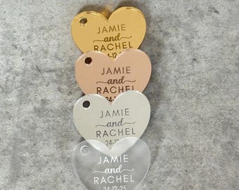 May include: Four heart-shaped acrylic tags in gold, rose gold, white, and frosted clear. Each tag has a hole for hanging and features the names "JAMIE and RACHEL" with a date below. The dates are 1-12, 24-, 24-10, and 24-12-25.