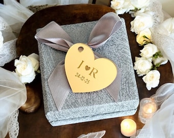Puede incluir: Una caja de regalo gris con una cinta plateada y una etiqueta dorada en forma de corazón. La etiqueta tiene las iniciales "J ❤️ R" y la fecha "24-12-25" grabadas. Rosas blancas y velas rodean la caja.