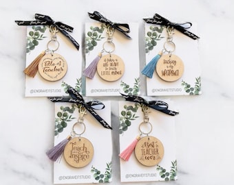 May include: Five wooden keychains with different teacher-themed sayings. Each keychain has a small tassel attached. The keychains are displayed on a white background with green foliage.