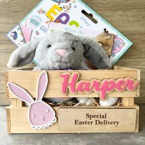 May include: A wooden crate with a pink bunny rabbit cutout, a grey plush bunny, and a pink wooden sign with the name "Harper" on it. The crate also has a sign that says "Special Easter Delivery".