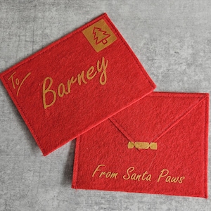 May include: Two red felt envelopes. One reads "To Barney" in gold script with a gold tree stamp. The other reads "From Santa Paws" in gold script with a gold clasp.