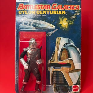 May include: A silver action figure of a Cylon Centurion from the Battlestar Galactica TV series. The figure is holding a gun and is packaged in a plastic blister pack with a colorful cardboard backing. The packaging features the show's logo and the text "Evil warrior from the TV Space Saga!"
