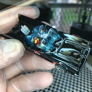 May include: A black die-cast toy car with a blue windshield and red accents. The car has a bat symbol on the side and two figures inside.
