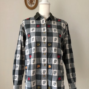 May include: A long-sleeved button-down shirt with a black, white, and gray checkered pattern. The shirt features embroidered leaf designs in red, yellow, and green. It has a green corduroy collar and wooden buttons.