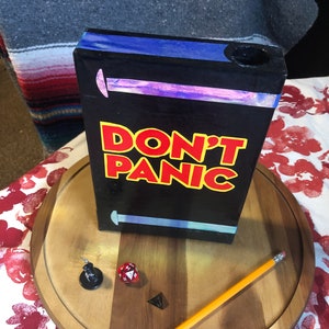 May include: A black wooden box with the words "DON'T PANIC" in yellow and red letters. The box has a blue and purple stripe on the top and a hole in the top. The box is sitting on a wooden round surface with a pencil, a red die, and a black miniature figure.