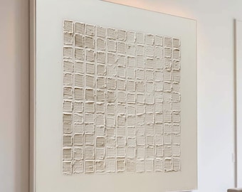 Puede incluir: Gran obra de arte abstracta cuadrada con un patrón texturizado en forma de cuadrícula en tonos beige y blanco. La obra está enmarcada en un borde blanco y cuelga de una pared blanca. La habitación cuenta con un sofá seccional de color claro, una otomana rectangular y una ventana grande.