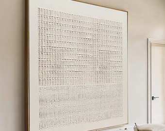 Puede incluir: Gran obra de arte abstracta en un marco dorado. La obra presenta un patrón similar a una cuadrícula en tonos beige y blanco. La habitación también incluye un sofá blanco, una mesa de centro de madera y dos otomanas.