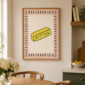 May include: A framed wall art print featuring a yellow stick of butter with the word BUTTER written in black on its side. The illustration is centered on a cream background, surrounded by a pink border with dark green dashed lines.