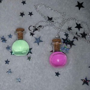 May include: Two glass potion bottle necklaces with cork stoppers. One bottle glows green, the other is pink. Both are suspended from silver chains. The background is a light gray with scattered star-shaped confetti.
