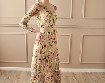 May include: A long, beige maxi dress with a floral embroidery pattern. The dress has a round neckline, long sleeves, and a fitted waist with a matching sash. The model is wearing the dress with gold heels.