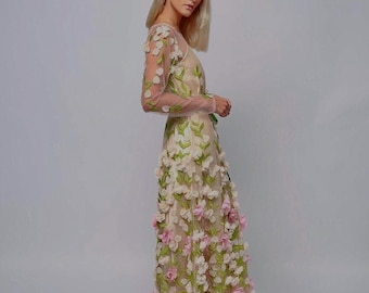May include: A floor-length, sheer gown adorned with three-dimensional floral appliqués in shades of pink, white, and green. The long-sleeved dress features a fitted bodice and a flowing skirt, creating an elegant silhouette.