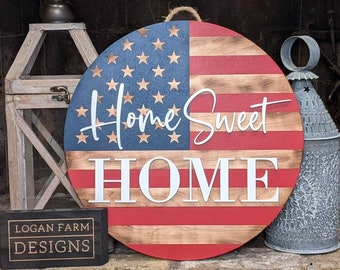 May include: A round wooden sign with a red, white, and blue American flag design. The sign says "Home Sweet Home" in white letters.