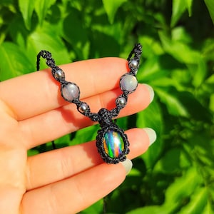 May include: A black macrame necklace featuring a central oval pendant with a dark, iridescent stone displaying vibrant flashes of green, orange, and blue. The cord is accented with two translucent round beads and four small metallic silver spheres.