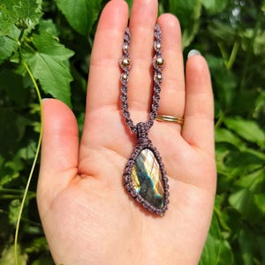 May include: A macrame necklace featuring a teardrop-shaped labradorite gemstone with iridescent blue and gold flashes. The stone is encased in dark grey knotted cord, which continues upward with integrated metallic beads in a symmetrical pattern.