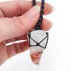 Puede incluir: Un colgante de cuarzo transparente con inclusiones rojas y naranjas, encerrado en una red tejida negra. El colgante cuelga de un cord&oacute;n negro con cuentas blancas y marrones. Un collar &uacute;nico, hecho a mano.