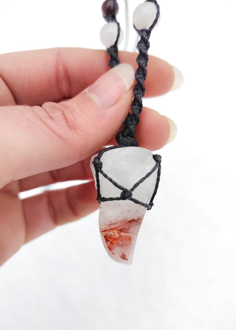 Puede incluir: Un colgante de cuarzo transparente con inclusiones rojas y naranjas, encerrado en una red tejida negra. El colgante cuelga de un cord&oacute;n negro con cuentas blancas y marrones. Un collar &uacute;nico, hecho a mano.
