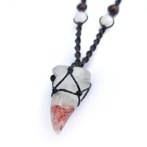 Puede incluir: Un collar con colgante de cristal transparente con un patr&oacute;n rojo y blanco. El cristal est&aacute; encerrado en un macram&eacute; negro tejido. El collar tiene un cord&oacute;n negro con cuentas blancas y marrones.