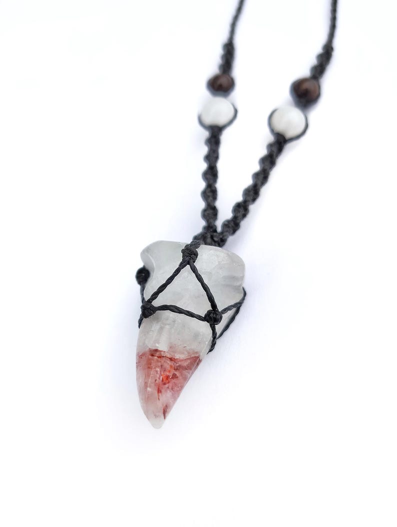 Puede incluir: Un collar con colgante de cristal transparente con un patr&oacute;n rojo y blanco. El cristal est&aacute; encerrado en un macram&eacute; negro tejido. El collar tiene un cord&oacute;n negro con cuentas blancas y marrones.