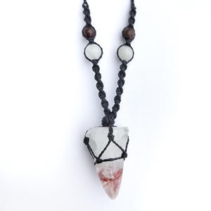 Puede incluir: Collar hecho a mano con un colgante de cristal de cuarzo transparente con tonos rojos y blancos, suspendido de un cord&oacute;n de macram&eacute; negro. El collar incluye cuentas marrones y blancas, creando una est&eacute;tica &uacute;nica. El colgante mide unos 5 cm de largo.