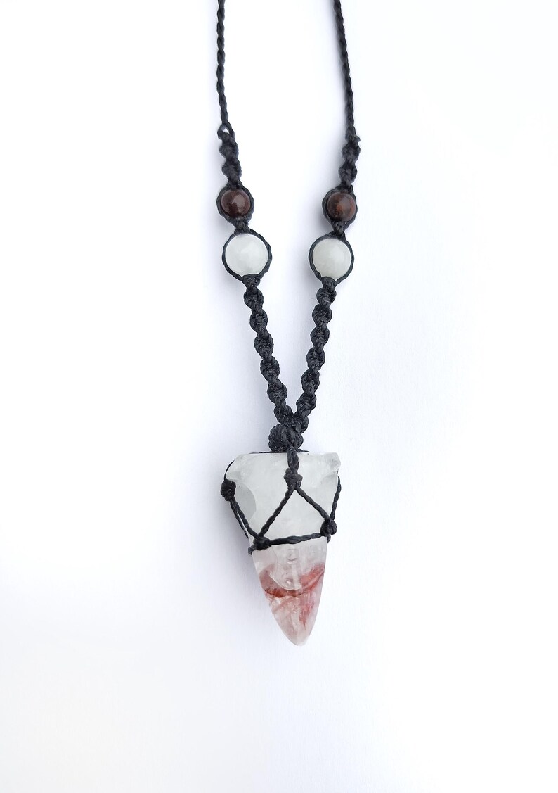 Puede incluir: Collar hecho a mano con un colgante de cristal de cuarzo transparente con tonos rojos y blancos, suspendido de un cord&oacute;n de macram&eacute; negro. El collar incluye cuentas marrones y blancas, creando una est&eacute;tica &uacute;nica. El colgante mide unos 5 cm de largo.