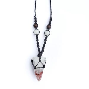 Puede incluir: Un collar hecho a mano con un colgante de cristal transparente con inclusiones rojas y blancas, suspendido de un cord&oacute;n de macram&eacute; negro. El cord&oacute;n est&aacute; adornado con cuentas marrones y blancas, creando una est&eacute;tica natural.