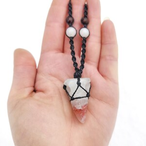 Puede incluir: Un collar hecho a mano con un colgante de cristal de cuarzo transparente con inclusiones rojas y blancas, envuelto en un macram&eacute; negro tejido. El collar tiene un cord&oacute;n negro con cuentas blancas y marrones.