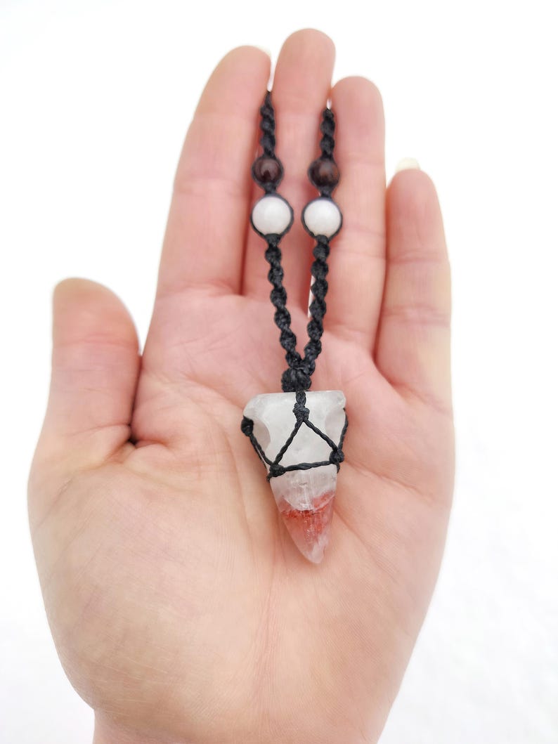 Puede incluir: Un collar hecho a mano con un colgante de cristal de cuarzo transparente con inclusiones rojas y blancas, envuelto en un macram&eacute; negro tejido. El collar tiene un cord&oacute;n negro con cuentas blancas y marrones.
