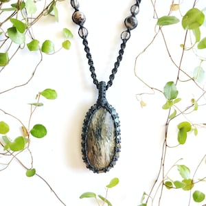 May include: A handcrafted black macrame necklace featuring a large, oval-shaped pendant with a natural stone center. The stone has a mix of brown, gray, and cream tones. The necklace also includes several smaller, round beads.