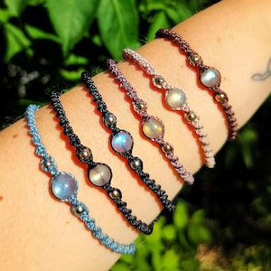 May include: Five macrame bracelets in blue, black, and brown tones, each featuring a central round iridescent gemstone flanked by two small metallic beads. The bracelets are constructed with knotted cord and arranged in a row on an arm.