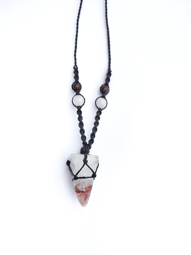 Puede incluir: Un collar hecho a mano con un colgante de cristal transparente con inclusiones rojas y blancas, suspendido de un cord&oacute;n de macram&eacute; negro. El cord&oacute;n est&aacute; adornado con cuentas marrones y blancas, creando una est&eacute;tica &uacute;nica y natural. El colgante mide aproximadamente 5 cm de largo.