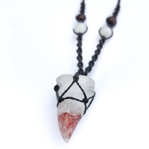 Puede incluir: Un collar con colgante de cristal transparente con un degradado rojo y blanco. El cristal est&aacute; encerrado en una red de macram&eacute; negro y ensartado en un cord&oacute;n negro con cuentas de madera y blancas. El collar se muestra sobre un fondo blanco.