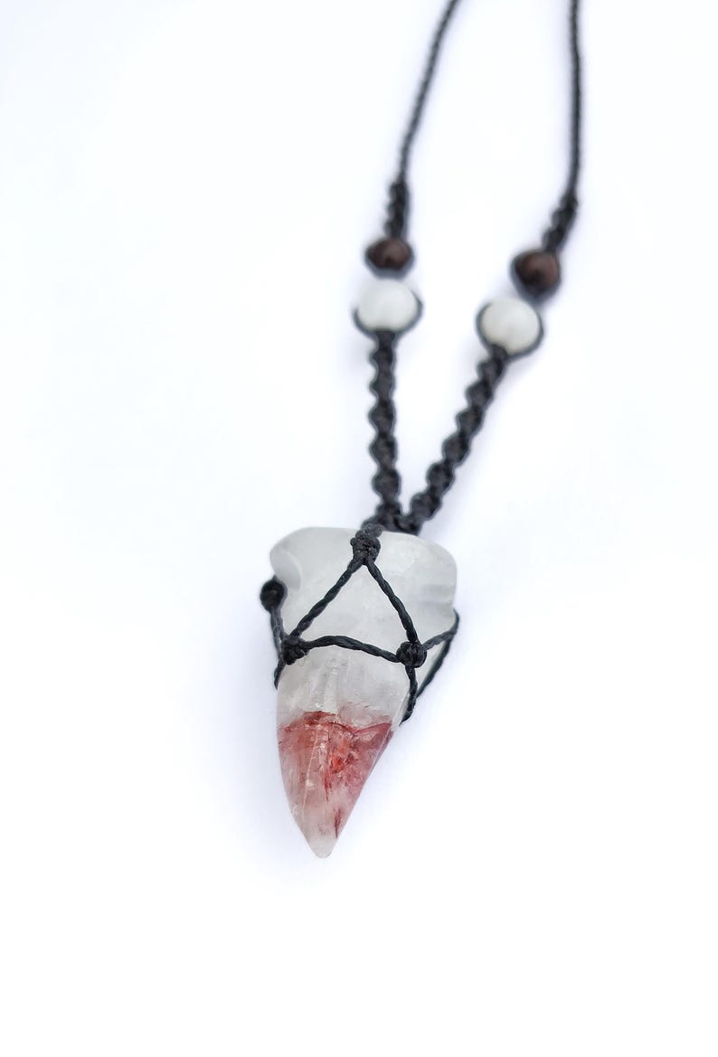 Puede incluir: Un collar con colgante de cristal transparente con un degradado rojo y blanco. El cristal est&aacute; encerrado en una red de macram&eacute; negro y ensartado en un cord&oacute;n negro con cuentas de madera y blancas. El collar se muestra sobre un fondo blanco.