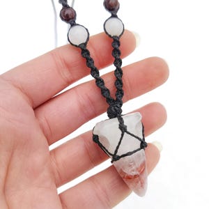 Puede incluir: Collar con colgante de cristal hecho a mano. El colgante es un cristal de cuarzo transparente con inclusiones rojas, suspendido en una red de macram&eacute; negra. El collar tiene un cord&oacute;n negro y cuentas blancas y marrones.