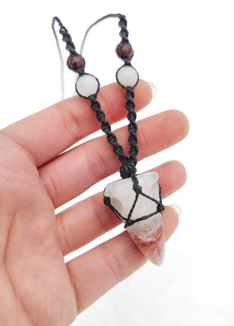 Puede incluir: Collar con colgante de cristal hecho a mano. El colgante es un cristal de cuarzo transparente con inclusiones rojas, suspendido en una red de macram&eacute; negra. El collar tiene un cord&oacute;n negro y cuentas blancas y marrones.