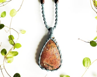 May include: A light blue macrame necklace with a pink and brown agate pendant. The necklace is photographed against a white background with green vines.