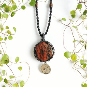 May include: A handcrafted necklace featuring a large, round, reddish-brown stone pendant with black veining. The pendant is suspended from a black macrame cord, accented with wooden beads. A silver-colored compass charm hangs below the stone.
