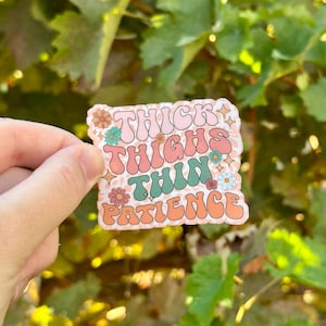 May include: A sticker with the text "Thick Thighs Thin Patience" in a retro font. The sticker has a peach background with floral accents in pink, green, and orange. The sticker is held against a blurred green background.