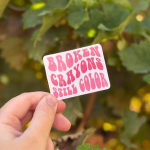 May include: A rectangular sticker with the text "BROKEN CRAYONS STILL COLOR" in pink, retro-style lettering. The sticker is held by a hand against a blurred background of green leaves and sunlight.