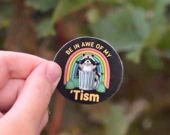 May include: A round sticker featuring a raccoon wearing sunglasses, sitting in a trash can, and a rainbow. The text on the sticker reads "BE IN AWE OF MY 'Tism". The sticker is black with a white border.