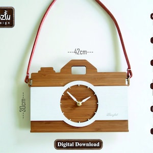 Puede incluir: Un reloj de pared de madera con forma de cámara, de 30 cm de alto y 42 cm de ancho, con un centro blanco y una esfera. El reloj tiene una correa roja para colgarlo. La esfera tiene manecillas doradas. Las palabras "toztu design" están en la esquina superior izquierda.