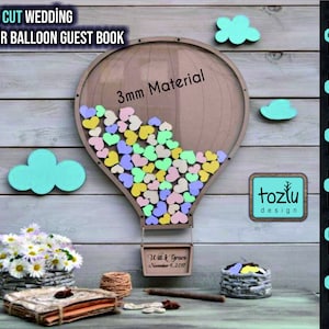 May include: Laser cut wooden hot air balloon guest book made from 3mm material. The balloon is filled with colourful hearts and has a basket at the bottom. The text "Will & Grace November 4, 2017" is written on a sign hanging from the basket. The image also includes a tozlu design logo.