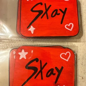 May include: Two red wooden squares with the name "Skay" written in black paint. Each square has white painted stars, hearts, and smiley faces.