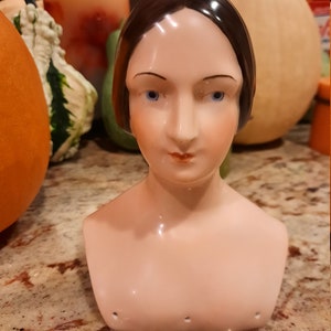 Könnte beinhalten: Eine Porzellanbüste einer Frau mit dunklen Haaren und blauen Augen. Die Büste ist realistisch bemalt und hat ein zartes, weibliches Aussehen.
